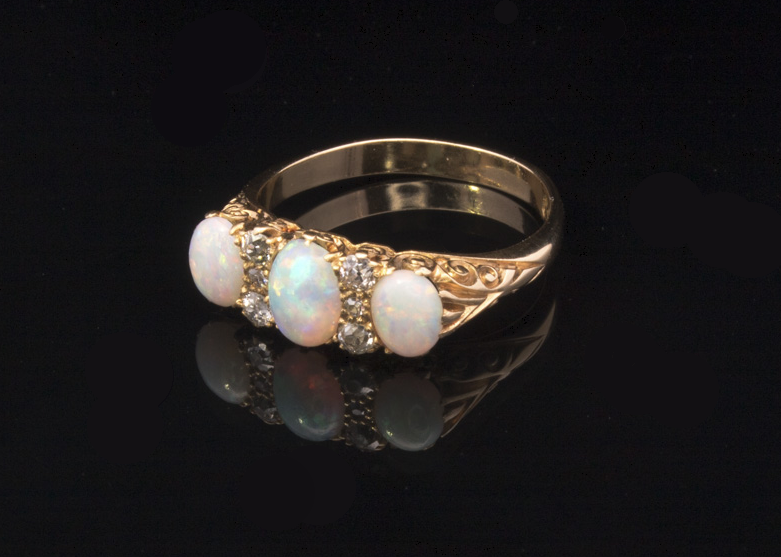 A yellow gold ring featuring three oval white opal gemstones and small clear accent stones, displayed on a reflective black surface.